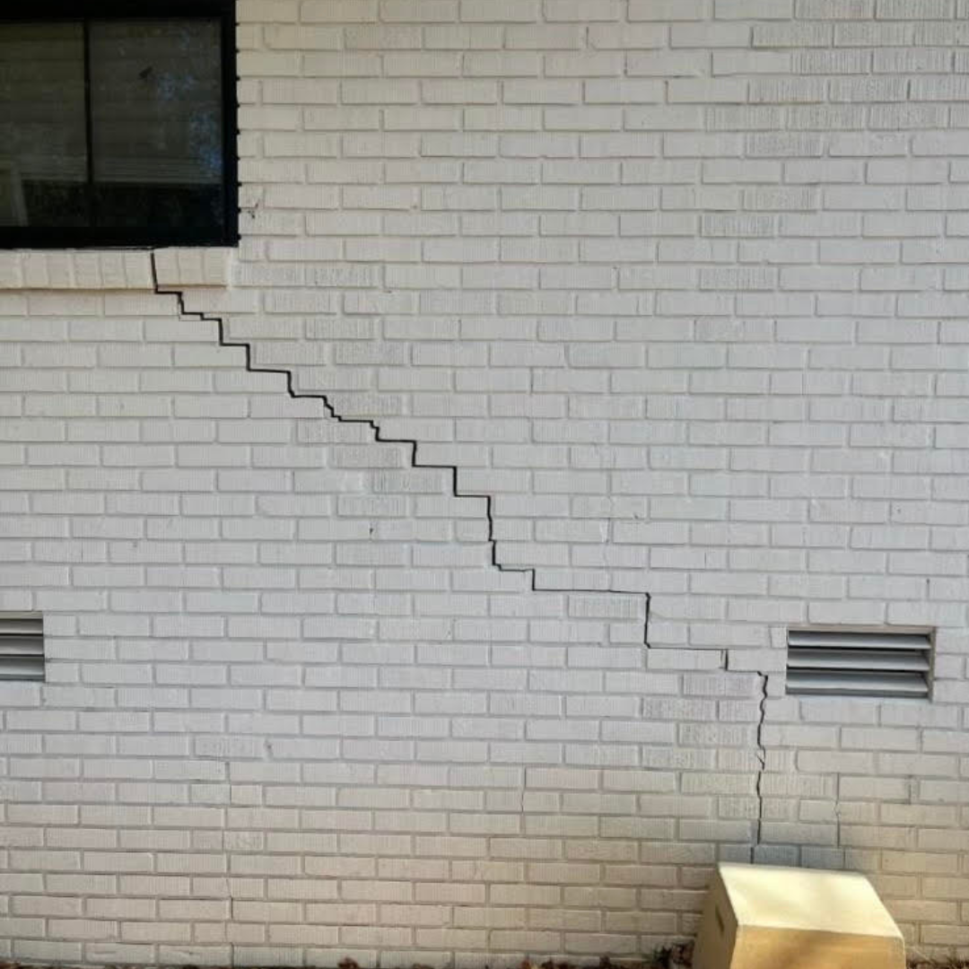 Stair-step crack in a white exterior brick wall, a common sign of foundation settlement and structural issues.